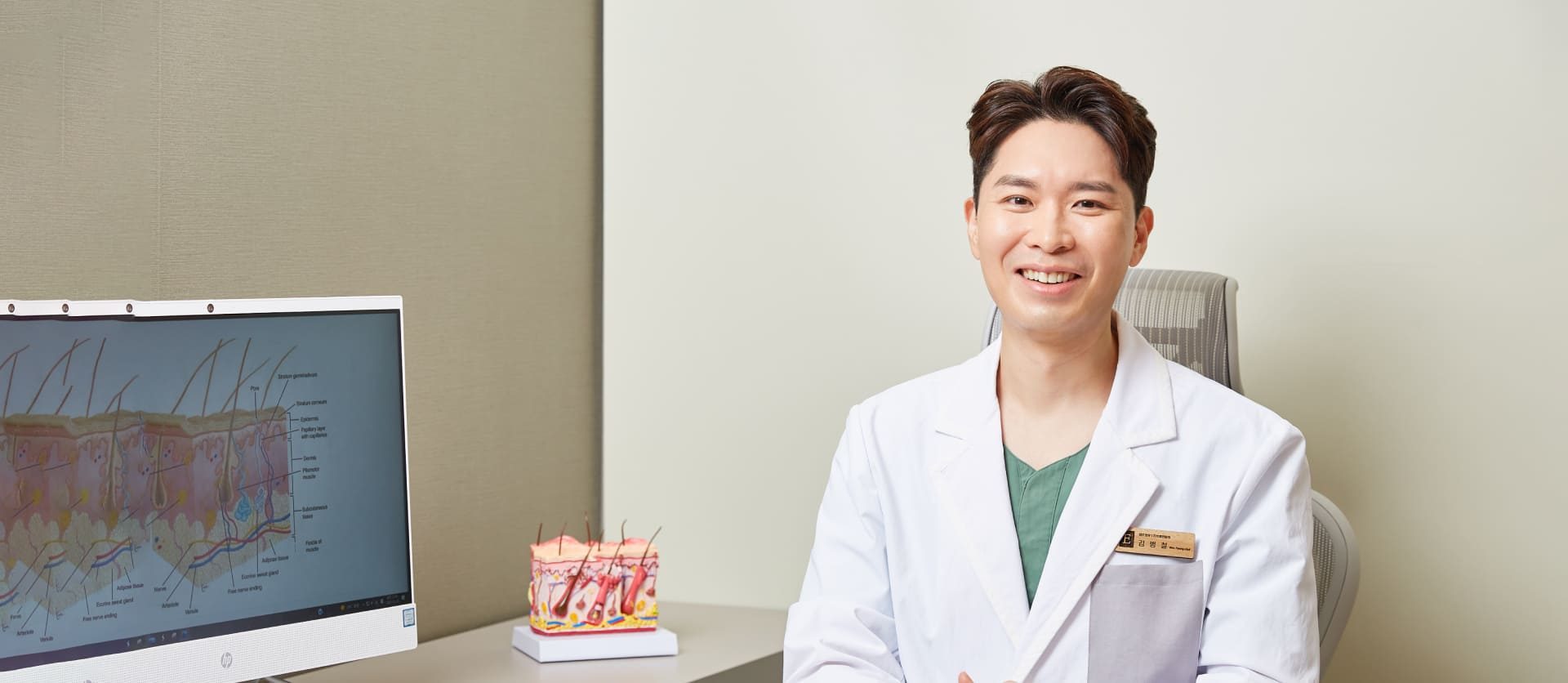 진료실 책상 앞에서 웃고 있는 김병철 대표원장(왼쪽에 피부 단면 그림 모니터와 피부 모형)